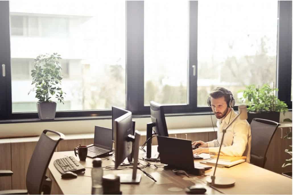 IT professional wearing a headset works at a desk with multiple computer monitors in a bright office.