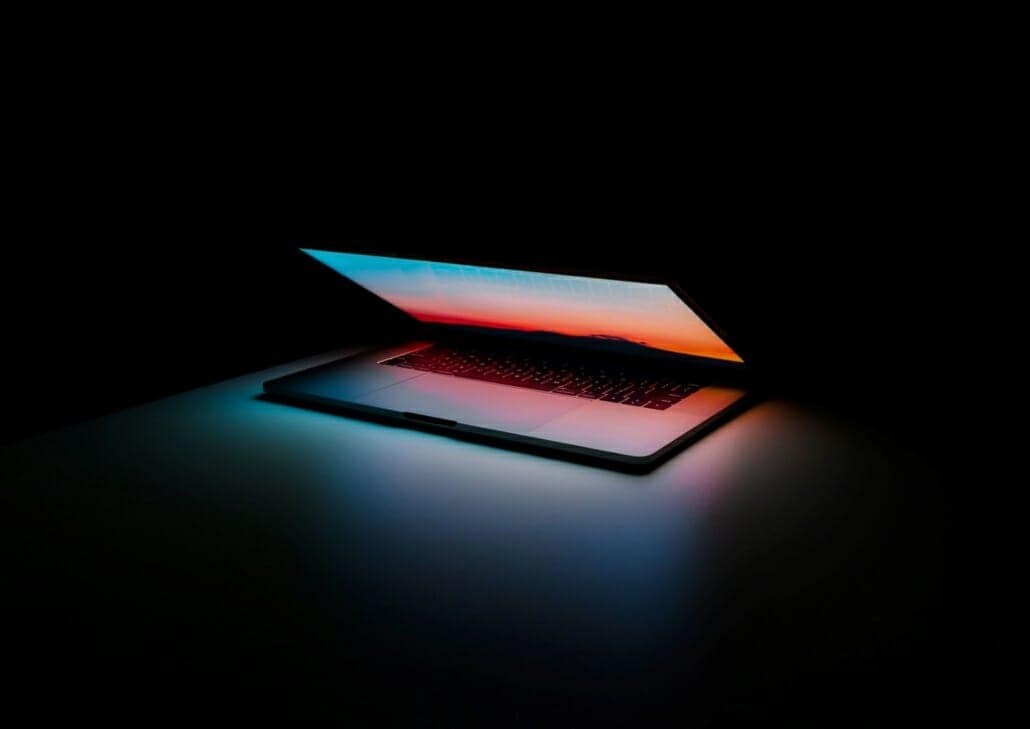 Laptop sitting in the dark with the lid open slightly, illuminating the room with the screen for cybersecurity trends to come.
