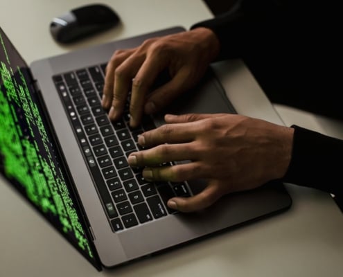 Close-up of hands typing on a laptop keyboard with green code displayed on the screen.