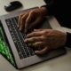 Close-up of hands typing on a laptop keyboard with green code displayed on the screen.