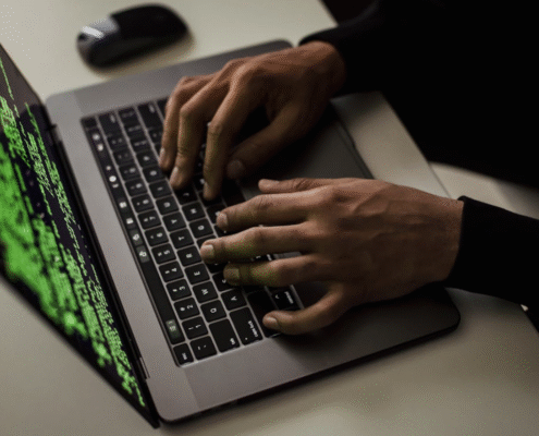 Hands typing on a laptop keyboard displaying green lines of computer code on the screen, viewed from an angled, close-up perspective on a desk with a computer mouse nearby.