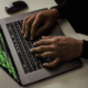 Hands typing on a laptop keyboard displaying green lines of computer code on the screen, viewed from an angled, close-up perspective on a desk with a computer mouse nearby.