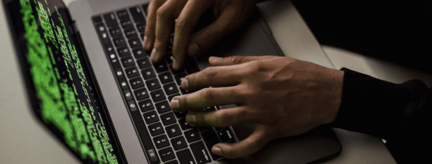 Hands typing on a laptop keyboard displaying green lines of computer code on the screen, viewed from an angled, close-up perspective on a desk with a computer mouse nearby.