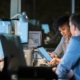 Two professionals working late in a modern office, reviewing information on a tablet beside computer monitors.