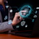 A business professional uses a fingerprint scanner on a digital interface beside a laptop, surrounded by cybersecurity, cloud, Wi-Fi, AI, and data icons.