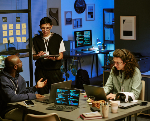 Three coworkers collaborate around laptops and a tablet in a modern office, with coding screens and project notes visible in the background.