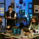Three coworkers collaborate around laptops and a tablet in a modern office, with coding screens and project notes visible in the background.
